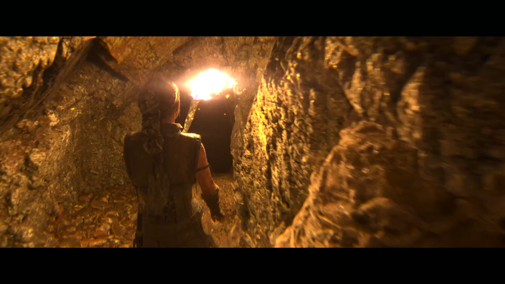 Senua walks through rocky cave toward bright opening